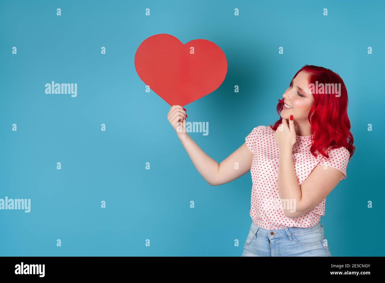 a dreamy contented young woman with red hair holds a red paper heart and scratches her chin with her hand , isolated on a blue background Stock Photo
