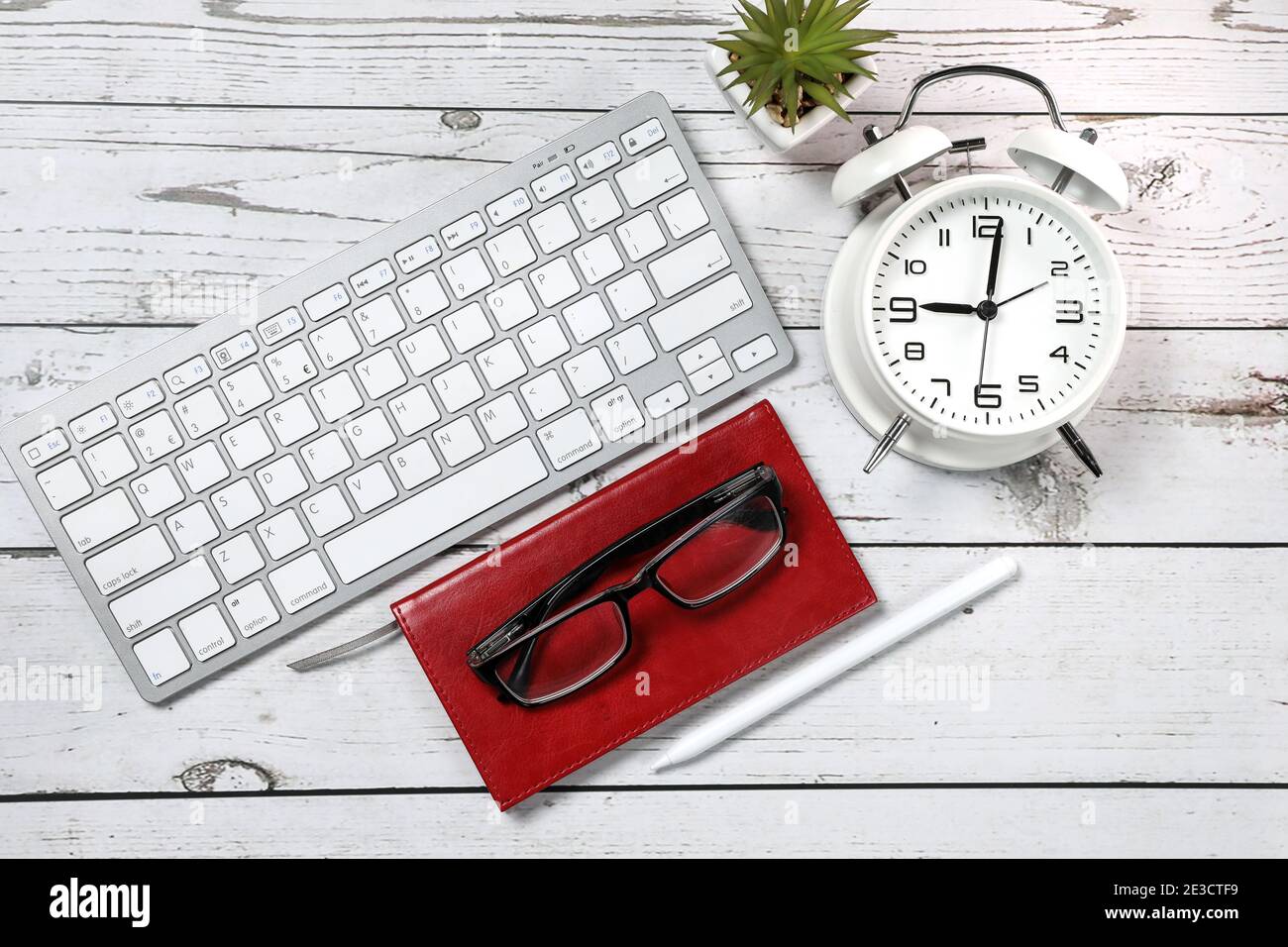 Flat lay view of a red notebook, white keyboard, pen and black glasses on a white wooden background. Top view of modern office workplace. Stock Photo