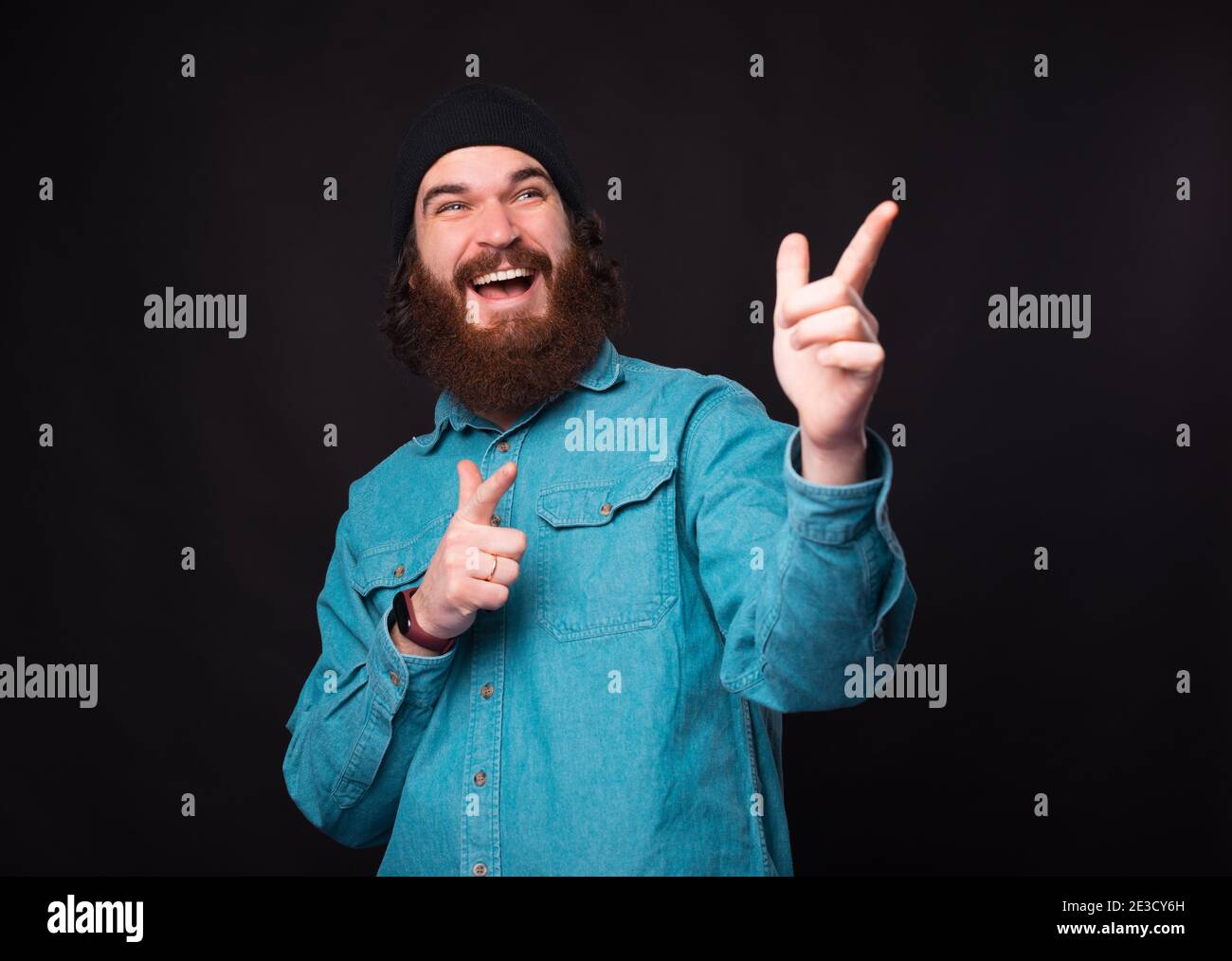 Cheerful bearded hipster man pointing away over dark background. Stock Photo