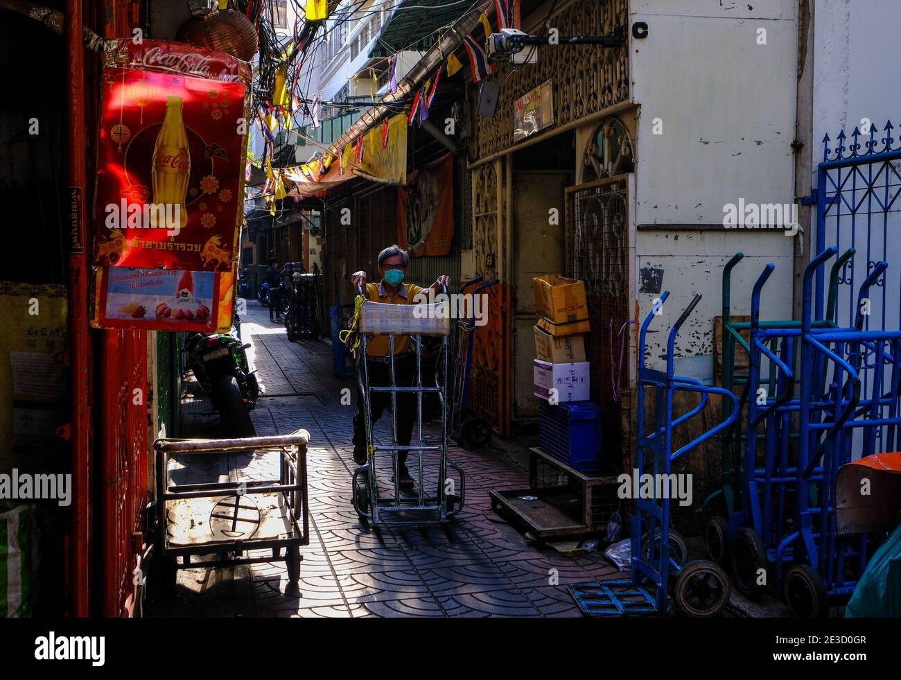 A man pushes an empty trolley along a sunlit road in Chinatown, Bangkok, Thailand Stock Photo