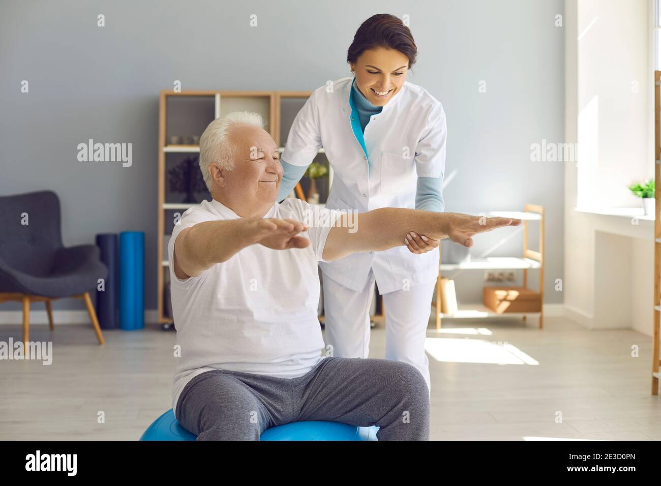 Happy senior man doing medical exercise on fitball with friendly nurse helping him Stock Photo