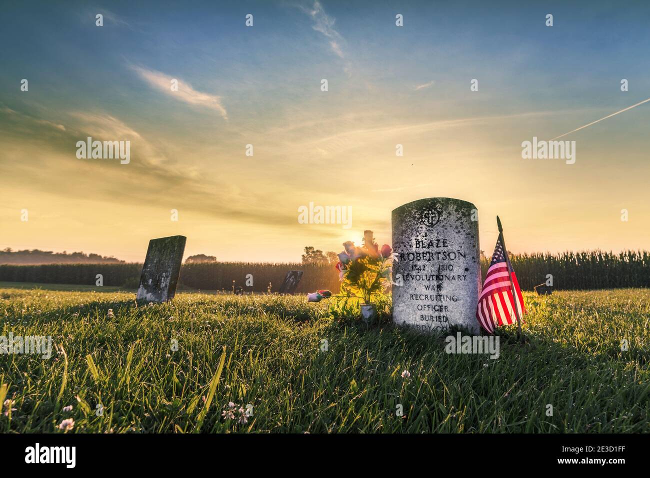 Sunrise over the headstone of a revolutionary war soldier on the Fourth of July.  The headstone is flanked by an American Flag and flowers. Stock Photo