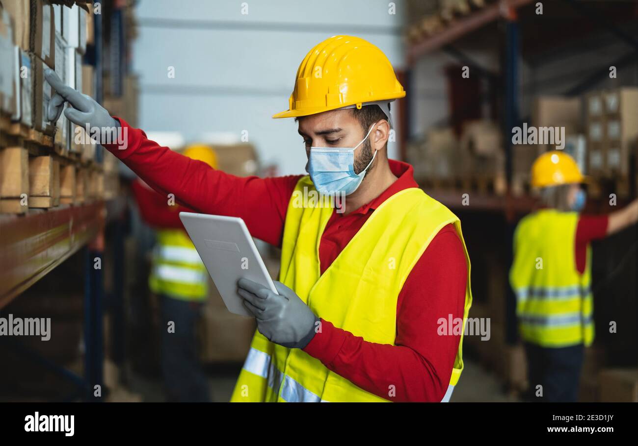 Young man working in warehouse doing inventory using digital tablet and loading delivery boxes plan while wearing face mask during corona virus Stock Photo