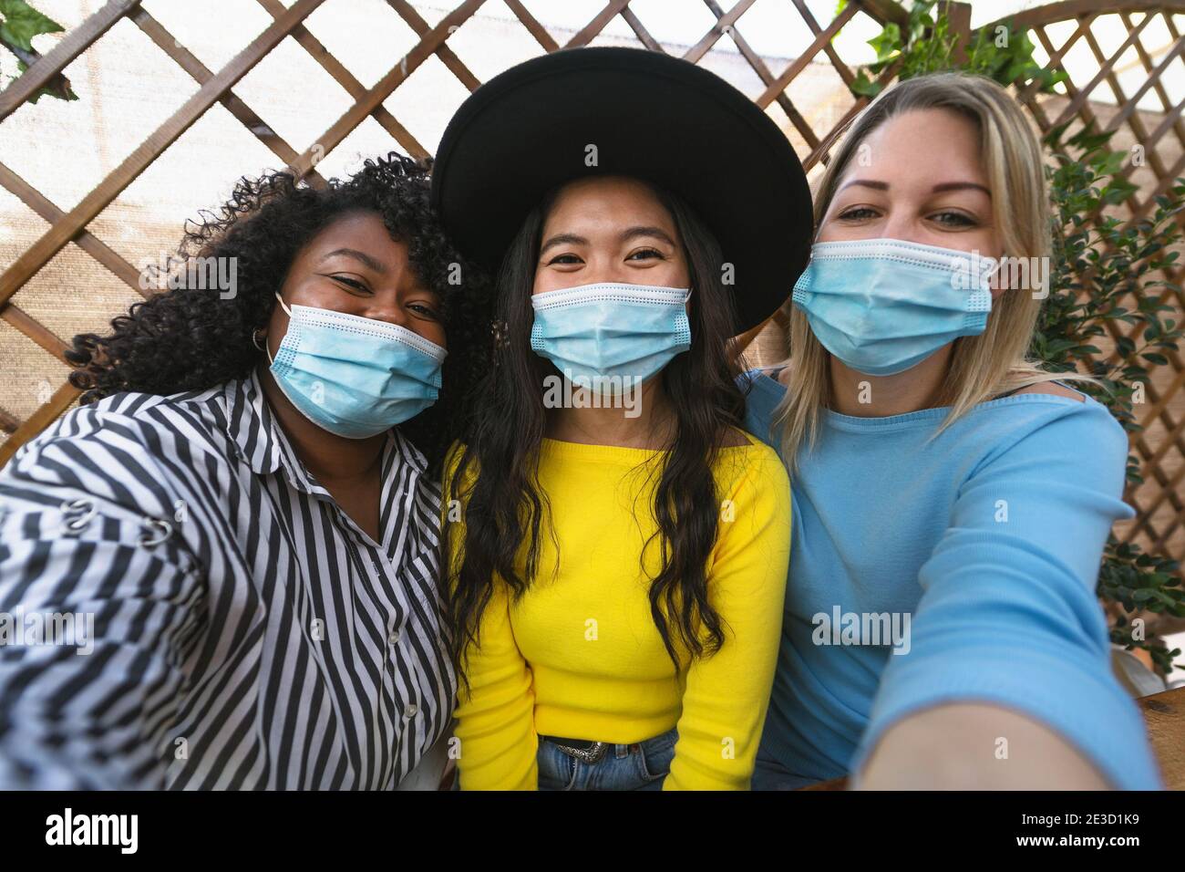 Multiracial friends wearing face mask while taking selfie with mobile smartphone cam during corona virus outbreak - Soft focus on central girl Stock Photo