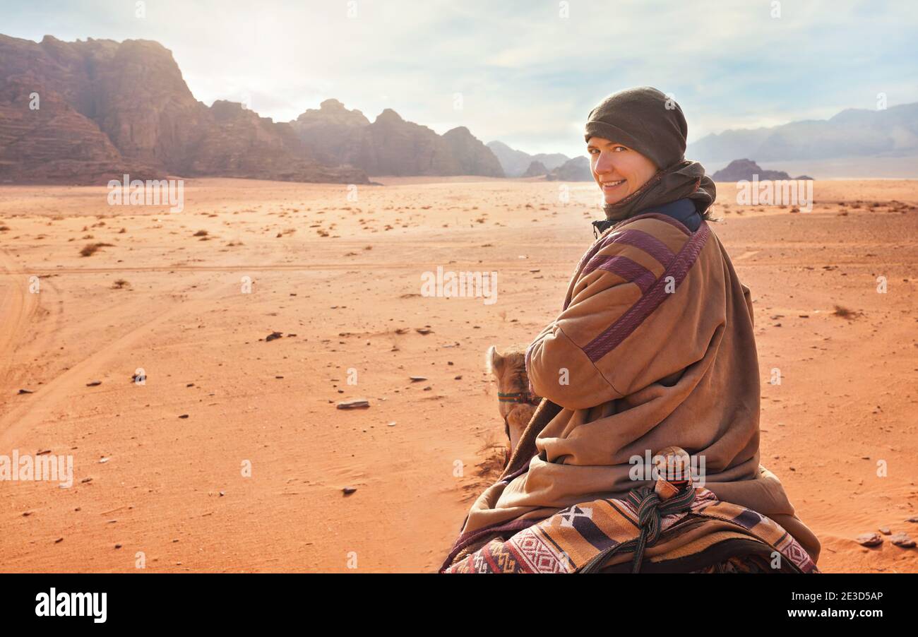 Young woman riding camel in desert, looking back over her shoulder, smiling. It's quite cold so she is wearing traditional Bedouin coat - bisht - and Stock Photo
