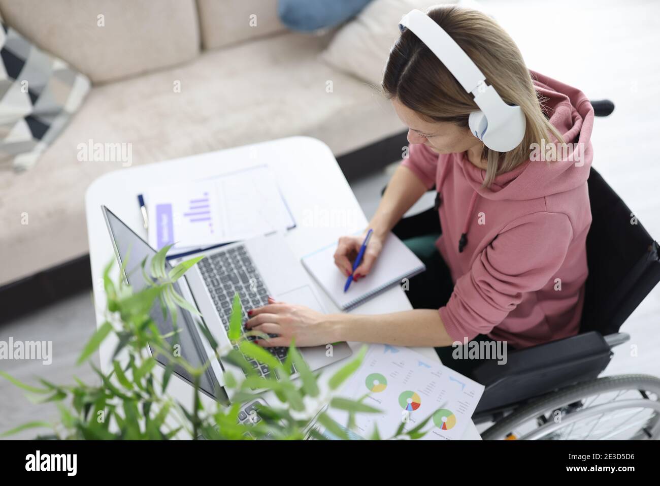 Woman in wheelchair sits at work desk wearing headphones and makes notes in notebook Stock Photo