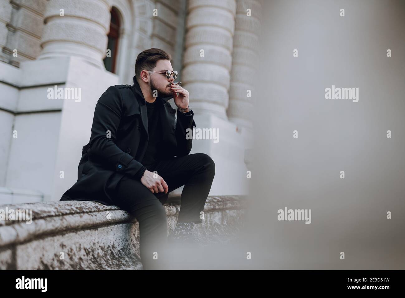 Calm young bearded guy in sunglasses smoking while sitting outdoors Stock Photo