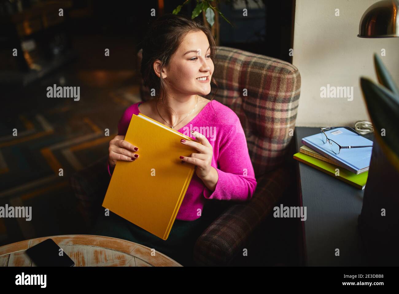 Smiling pretty lady sitting in cozy armchair and looking out the window in cafeteria Stock Photo