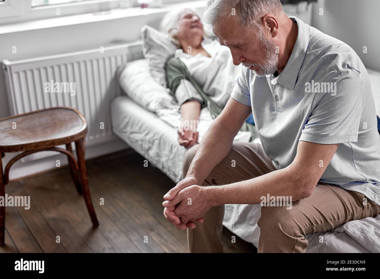 depressed man sits near his sick elderly wife lying on bed suffering from disease. in hospital Stock Photo