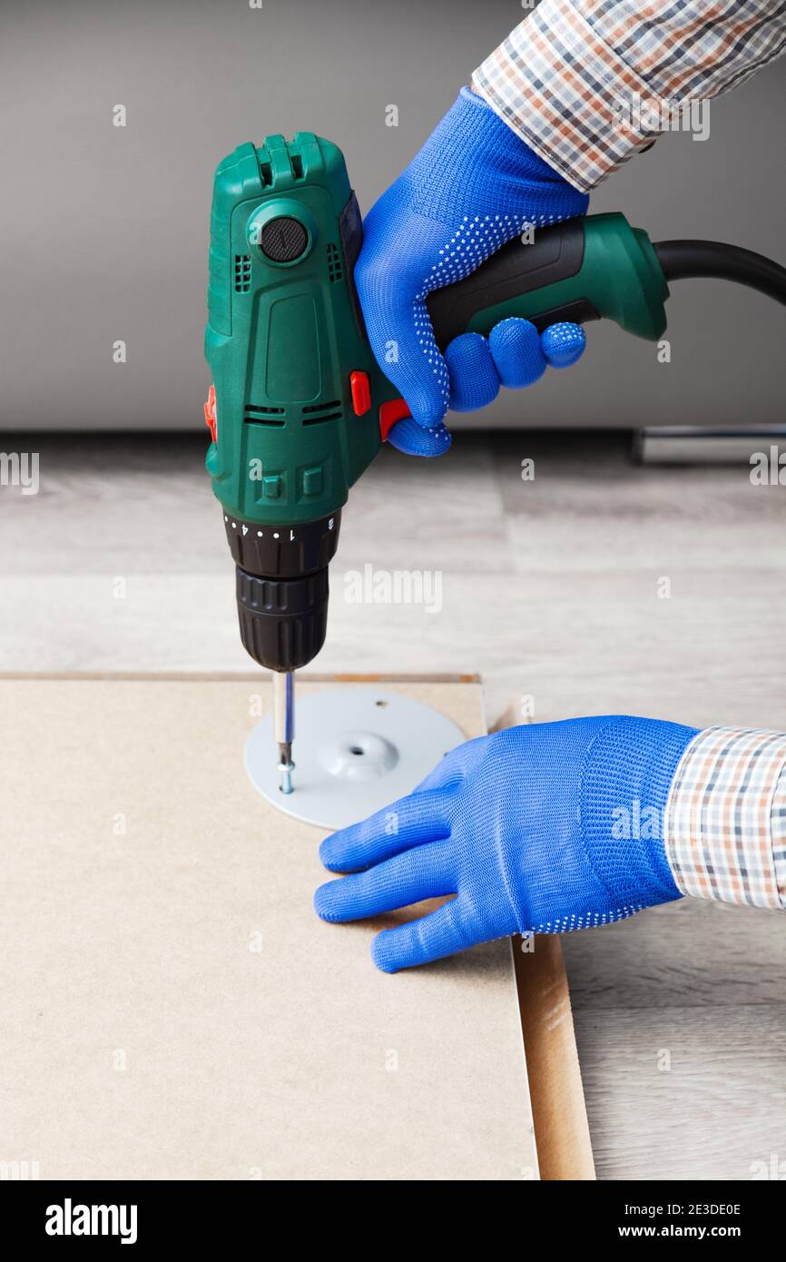Electric Drill works in handyman hands at home. Male hands in gloves using electric drill tool to assemble and repair furniture. Home DIY Stock Photo