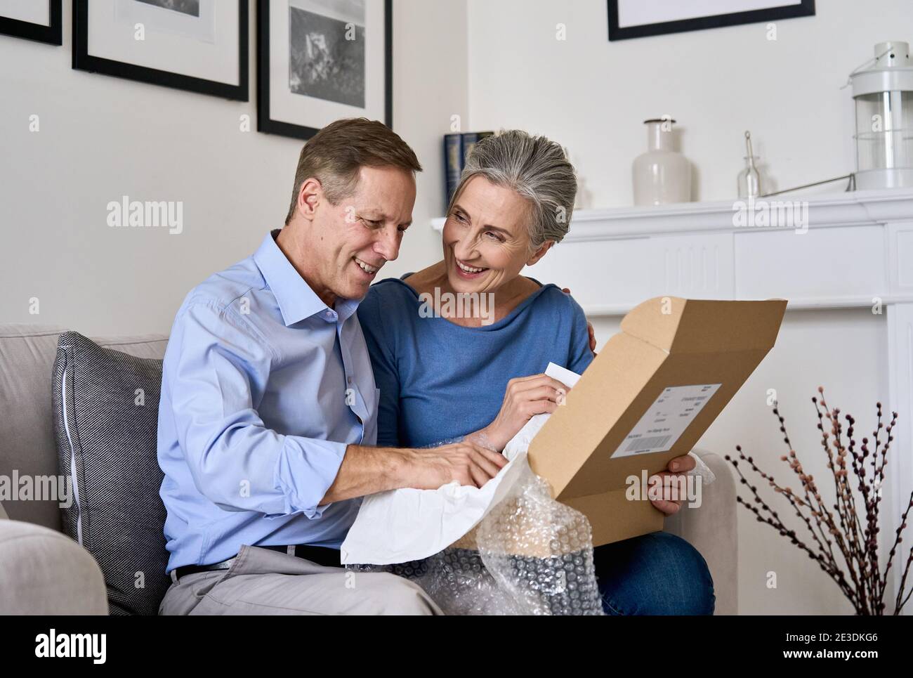 Happy old senior couple unpacking shipping delivery box sitting at home on sofa. Stock Photo