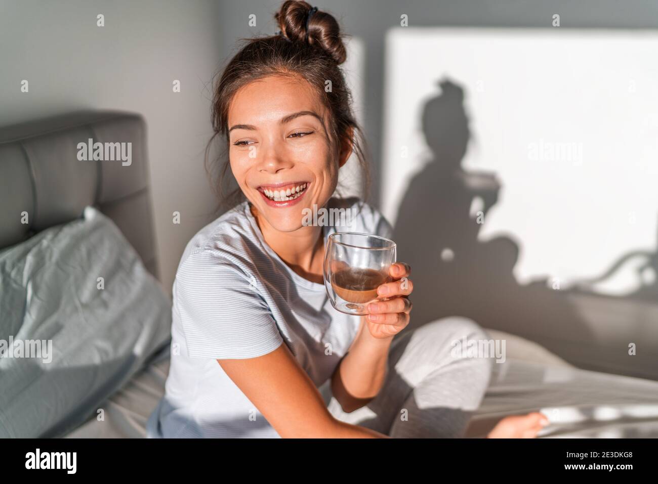 Happy morning Asian woman drinking coffee cup in bed happy chinese casual girl waking up with hot beverage mug relaxing smiling on comfy mattress Stock Photo