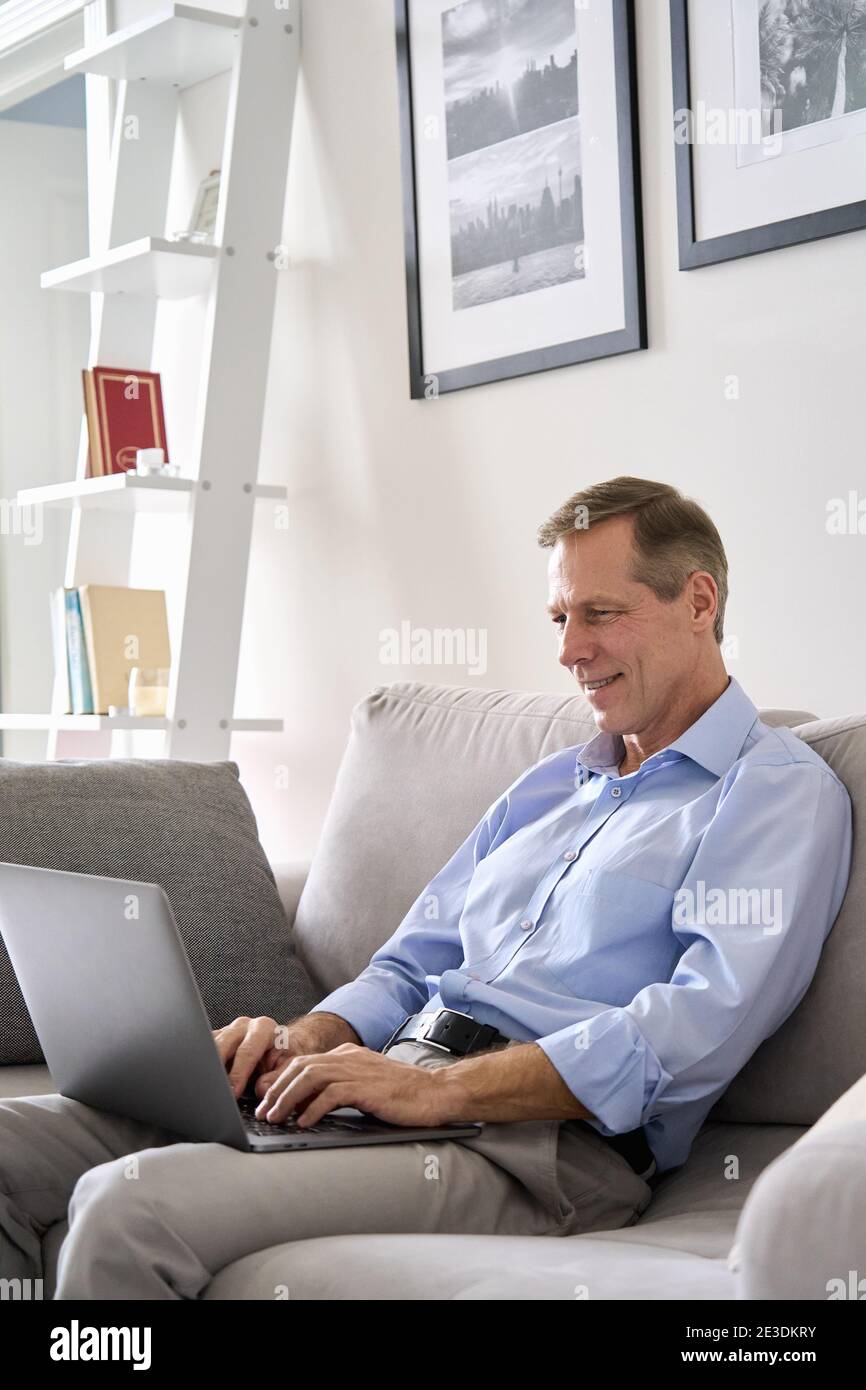 Smiling mature man typing on laptop sitting on sofa working online at home. Stock Photo