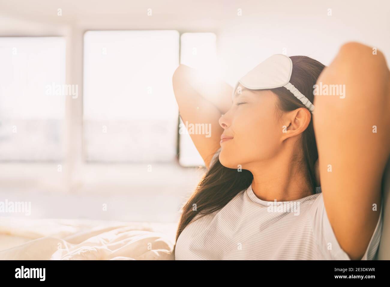 Sleeping at home woman waking up in morning sunlight stretching happy after a good night sleep feeling rested wearing eye mask. Asian girl bed sleep Stock Photo