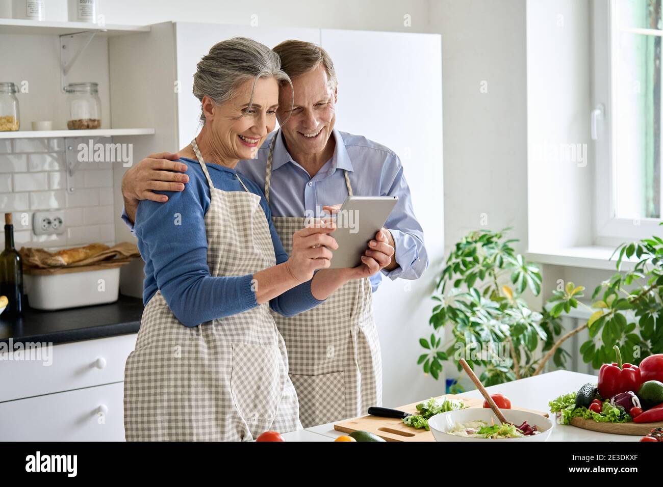 Happy middle aged couple preparing salad using tablet in kitchen. Stock Photo