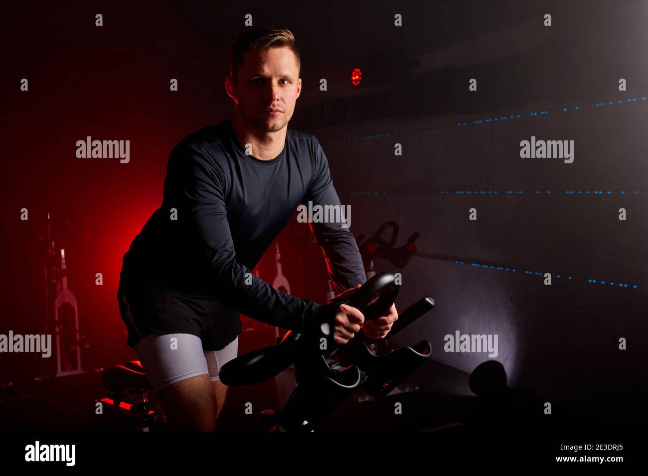 sportsman training at sport equipment in the gym, confident and strong male in tracksuit has intensive training on bicycle in fitness club, looks at c Stock Photo