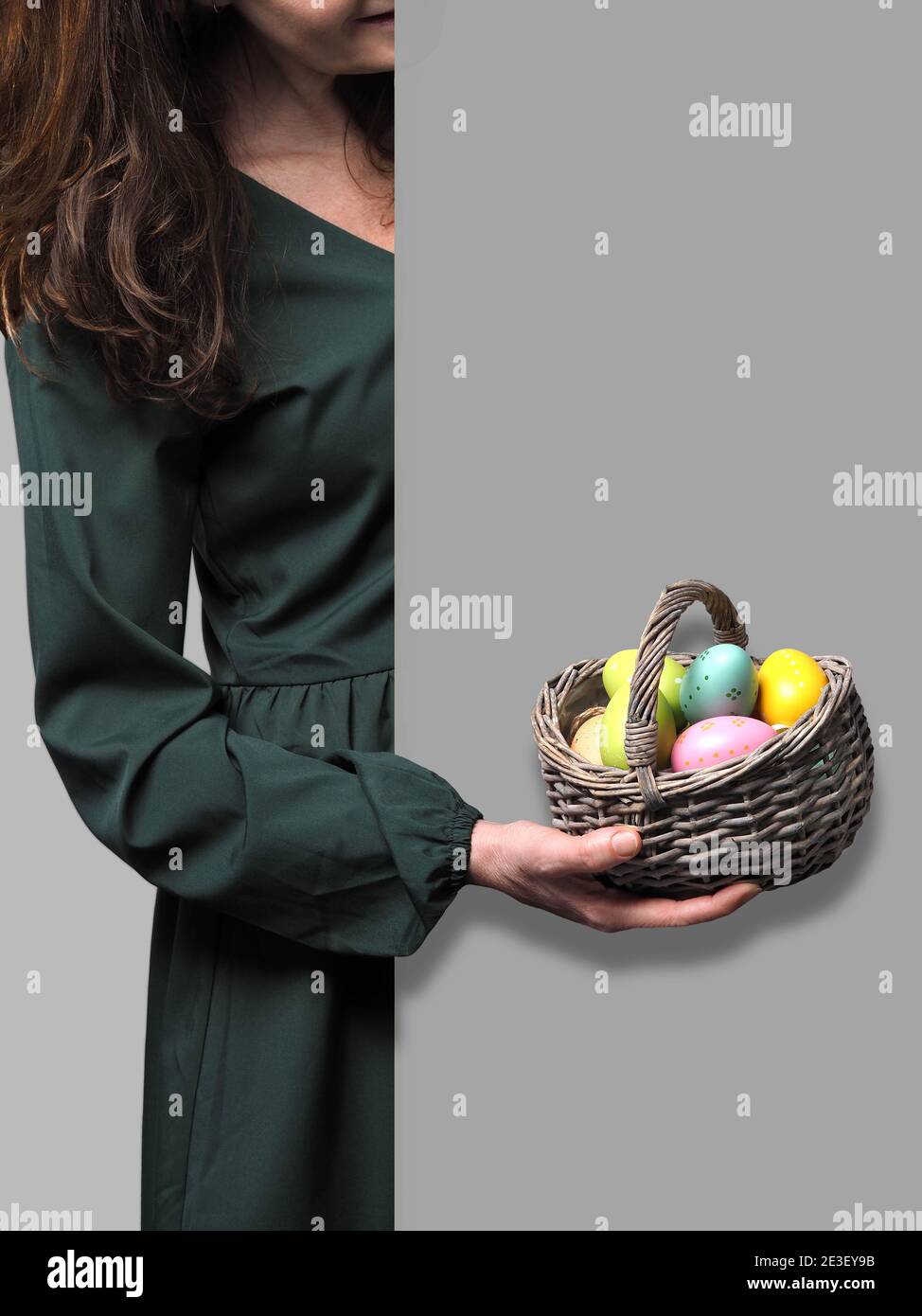 Female hand in green dress holding basket with colored decorative easter eggs in front of grey wall. Copy space.  Stock Photo