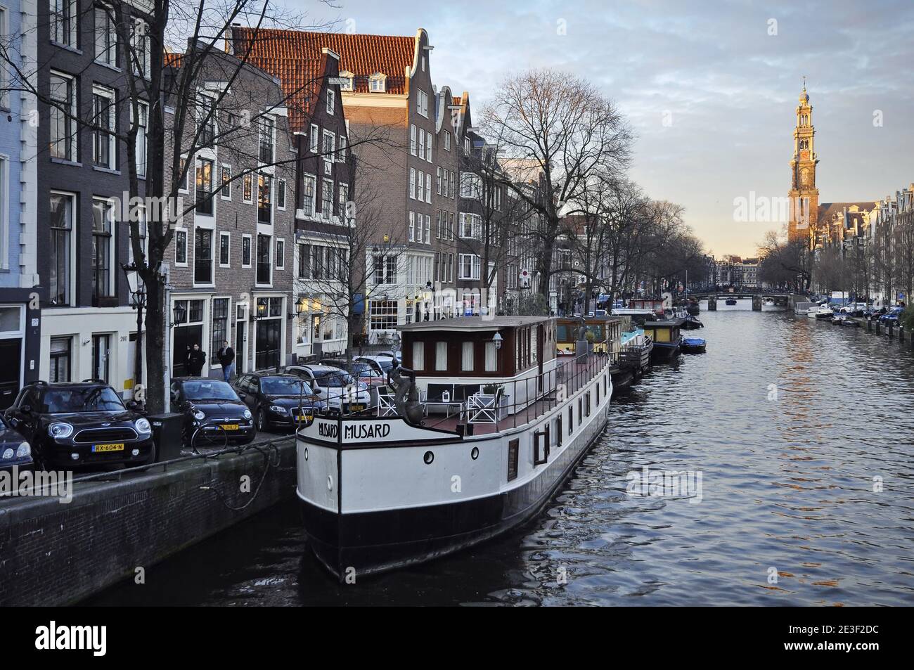 A white house boat named Musard is docked on the left side of the canal. The Westerkerk church is in the background, under the afternoon sunshine Stock Photo