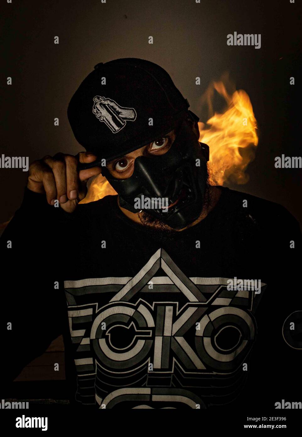 Man wearing a black mask and a cap with a graffiti spray can on it with a fire in the background Stock Photo