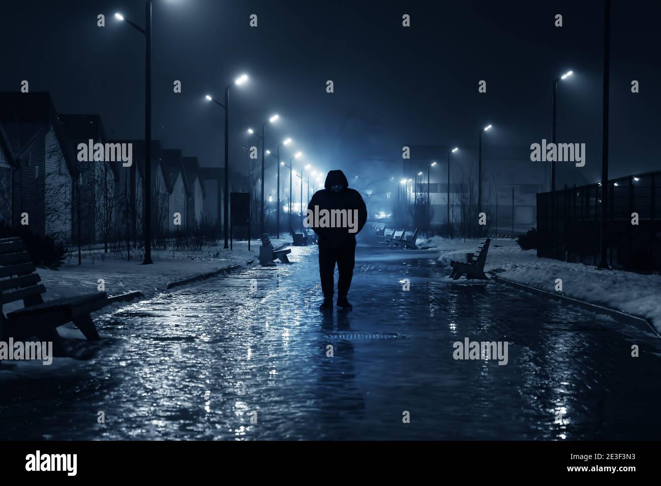Silhouette of lonely person walks on dark foggy street illuminated with street lamps, blue toned. Stock Photo