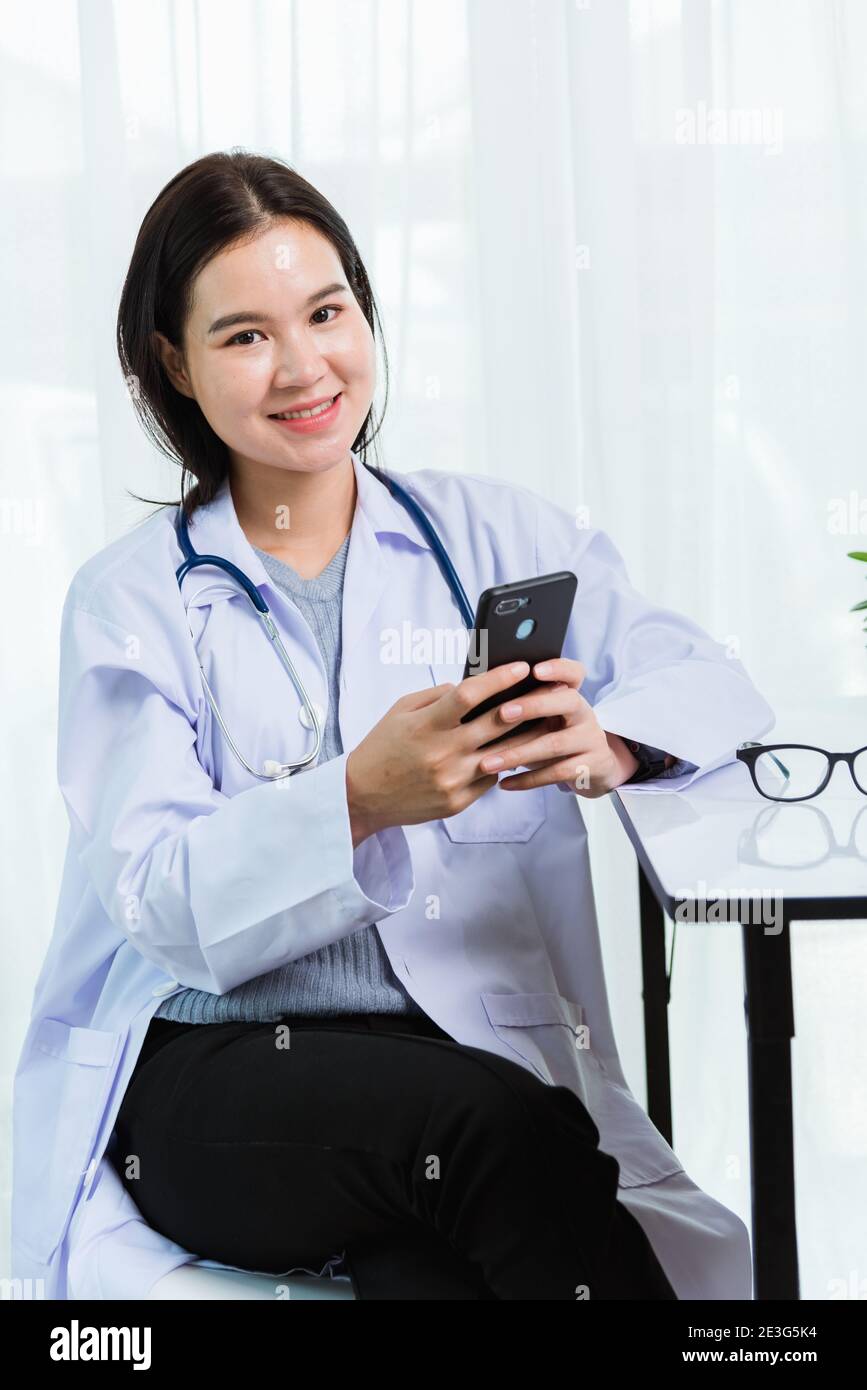 Asian doctor young beautiful woman smiling using working or holding with smart mobile phone and laptop computer at hospital desk office, technology he Stock Photo