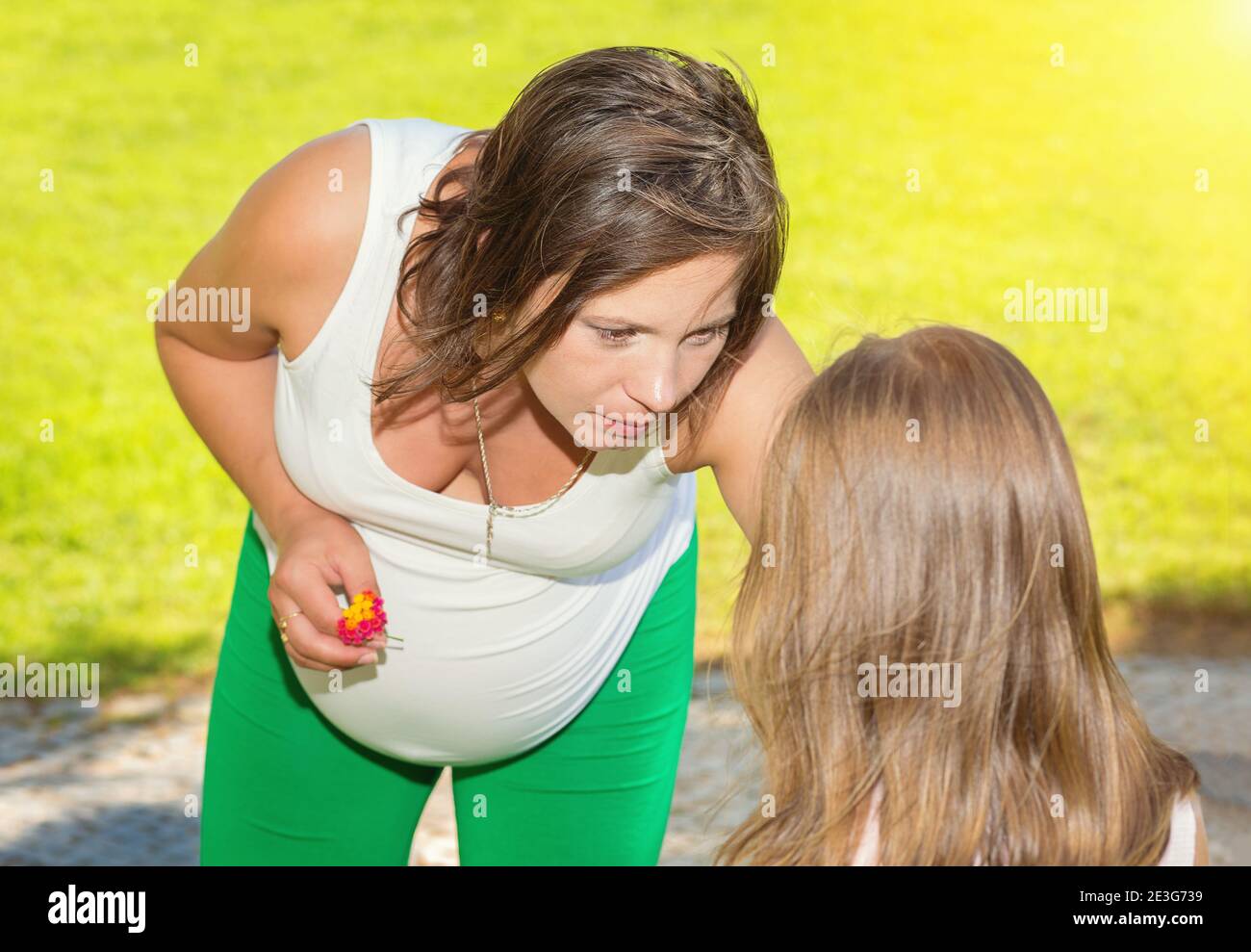 Pregnant woman explaining something to her daughter, girl who is jealous on her mother about the new pregnancy sitting outdoors on a green grass meado Stock Photo