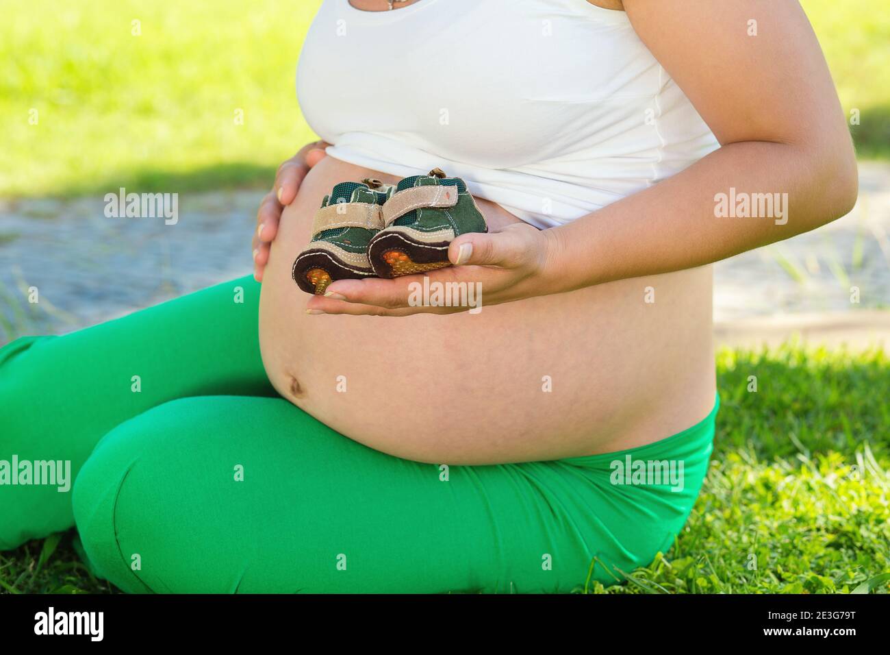 Unrecognnizable Pregnant Woman touching her belly, tummy playing with small baby boy shoes sitting outdoors on a green grass meadow in a park on sunny Stock Photo
