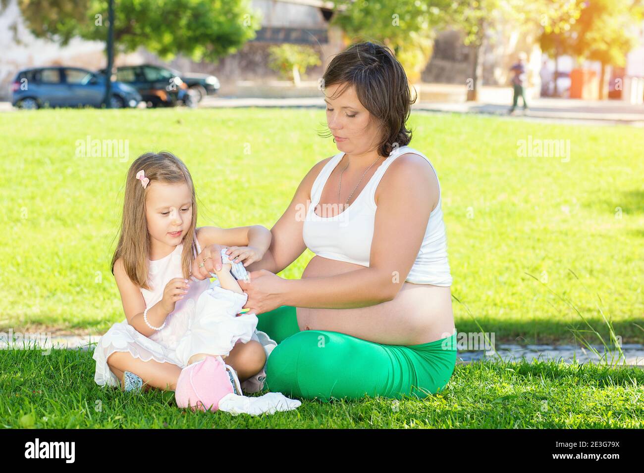 Kid girl withpregnant mom resting playing with a doll in park on green grass. Girls playing with toys. Both and wearing white clothing dresses. Picnic Stock Photo
