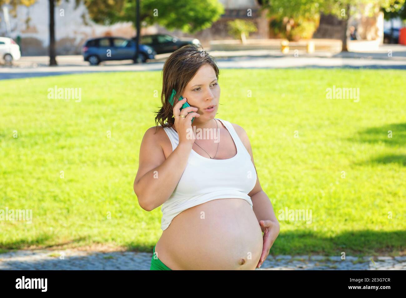 Emotional pregnant woman talking by mobile phone angry about to deliver outdoors while sitting on green grass meadow. Emergency call Stock Photo
