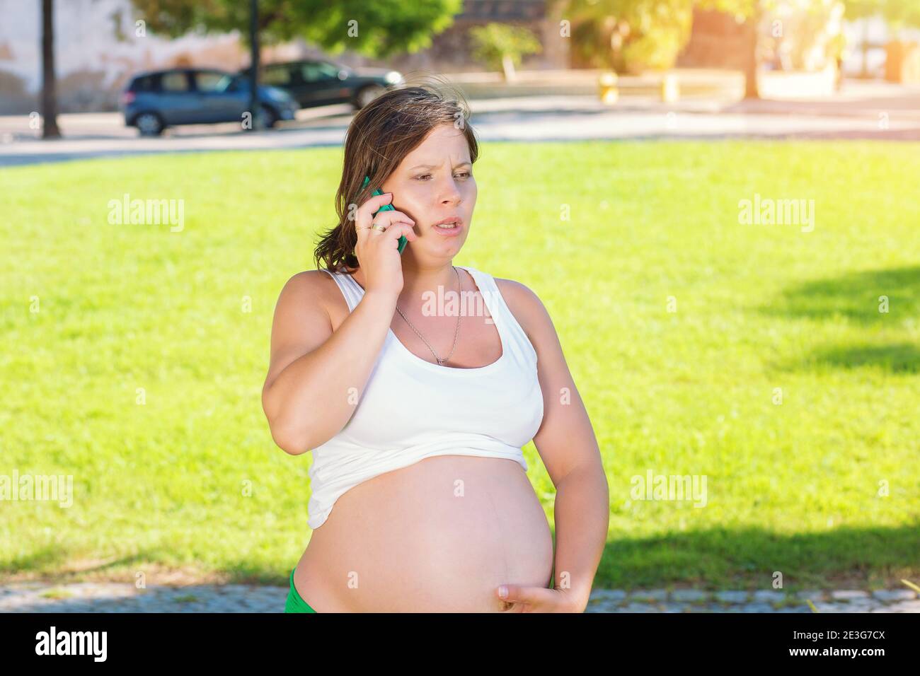 Emotional pregnant woman talking by mobile phone angry about to deliver outdoors while sitting on green grass meadow. Emergency call Stock Photo