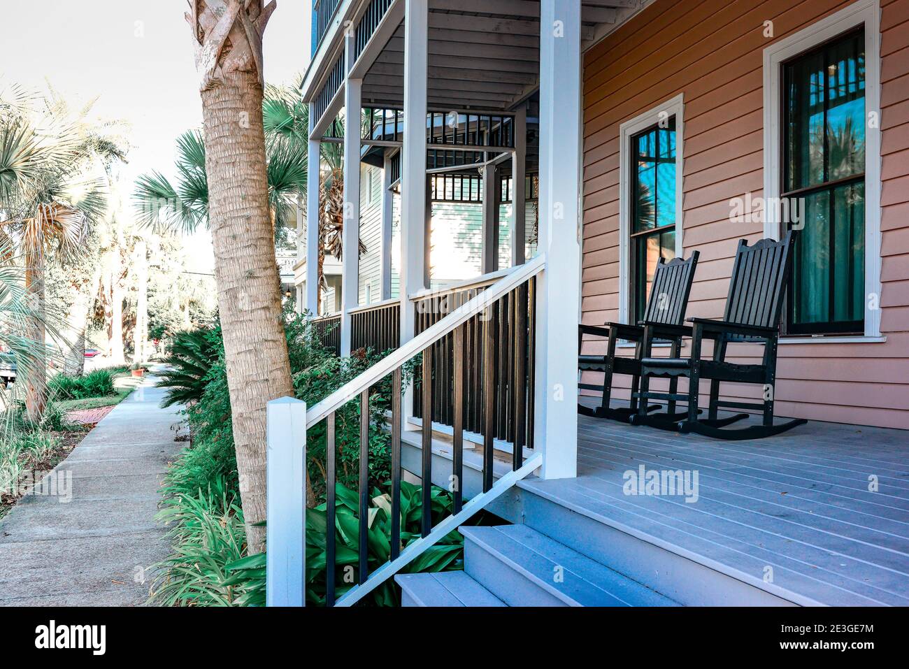 Rocking chairs on the front porch of a two-storied Inn with vanishing sidewalk into sunlight drenched palm trees in Fernandina Beach, FL Stock Photo