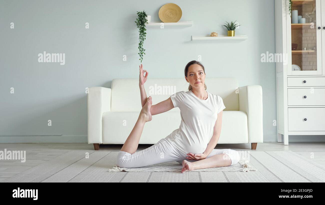 Calm young pregnant brunette woman does ardha matsyendrasana practicing yoga position on floor near sofa in spacious room at home Stock Photo