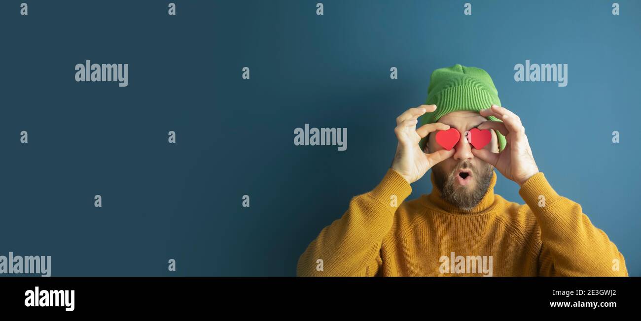 Portrait of a cheerful funny young hipster man with hearts in his eyes on a blue background. Sales, discounts, Valentine's Day and love concept. Stock Photo