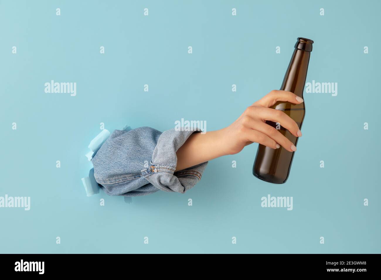 Female hand with bottle of beer breaks through blue paper background