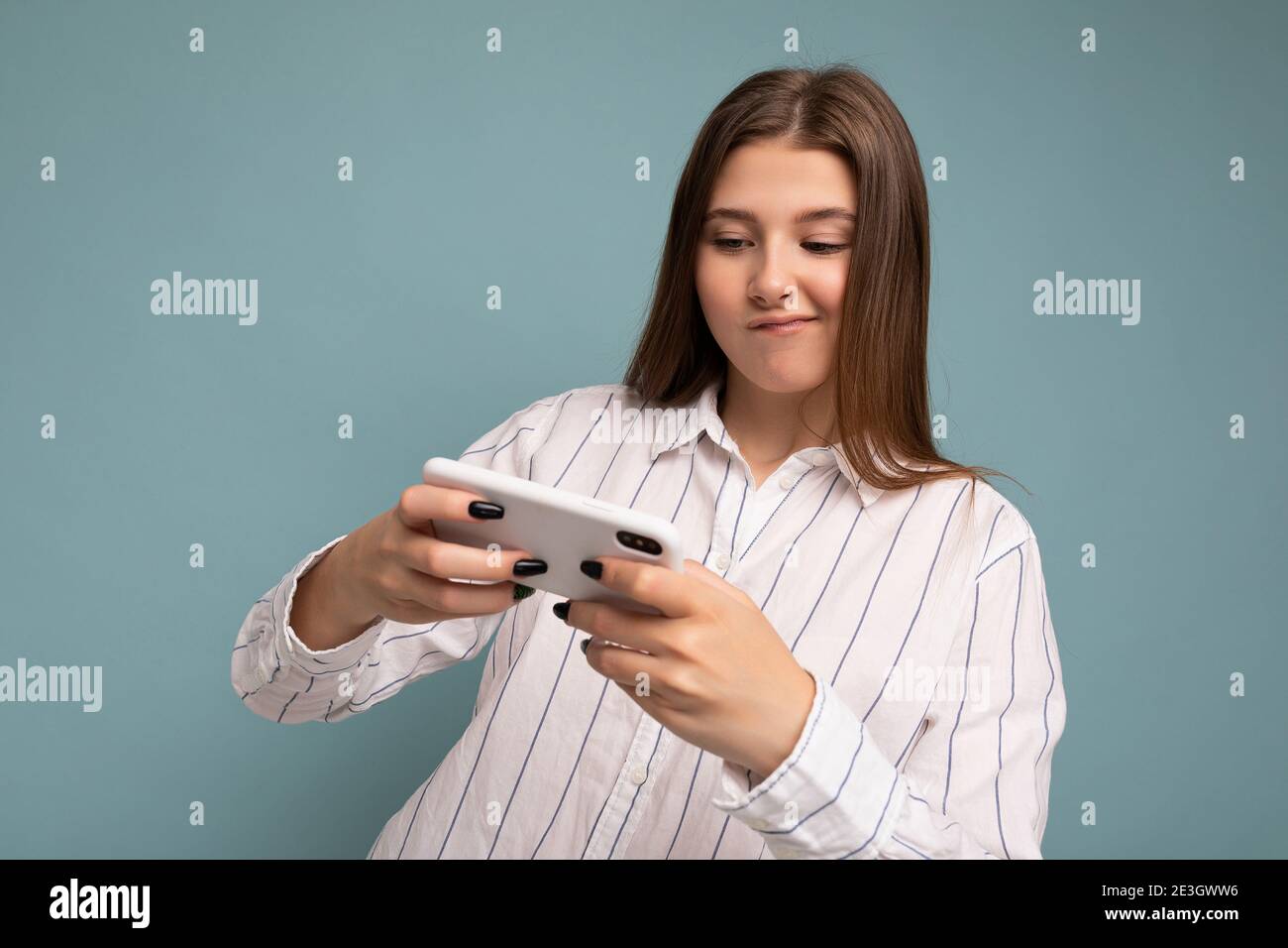 Concentrated happy attractive young blonde woman wearing casual white shirt isolated over blue background wall holding smartphone and playing online Stock Photo