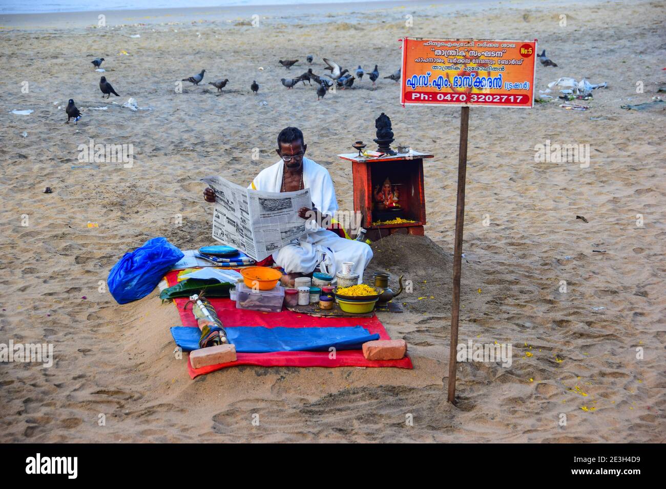 Hindu Priest reading newspaper, before washing away sins of pilgrims, Varkala Beach, Varkala, Kerala, India Stock Photo