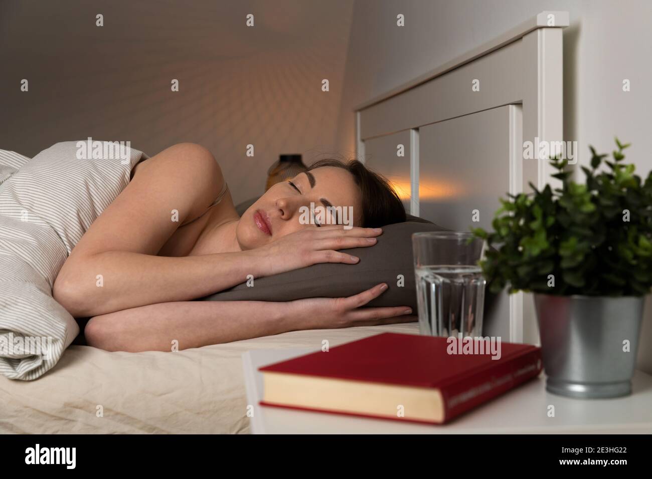 Young women sleeping in bed with a warm light in background and a bedside table in front with clear drinking water and a book. Stock Photo