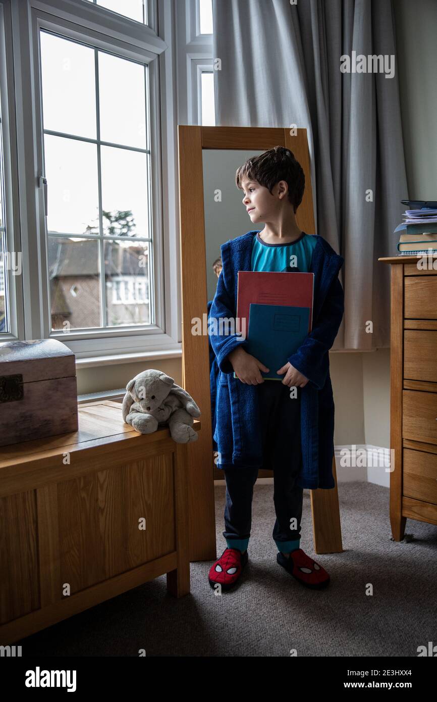 7 year old boy looks out of the bedroom window thinking of his school friend as he holds his school books whilst wearing pyjamas and dressing gown UK Stock Photo