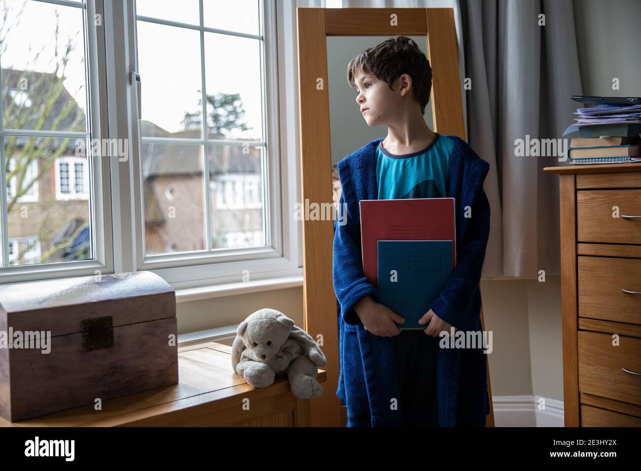 7 year old boy looks out of the bedroom window thinking of his school friend as he holds his school books whilst wearing pyjamas and dressing gown UK Stock Photo