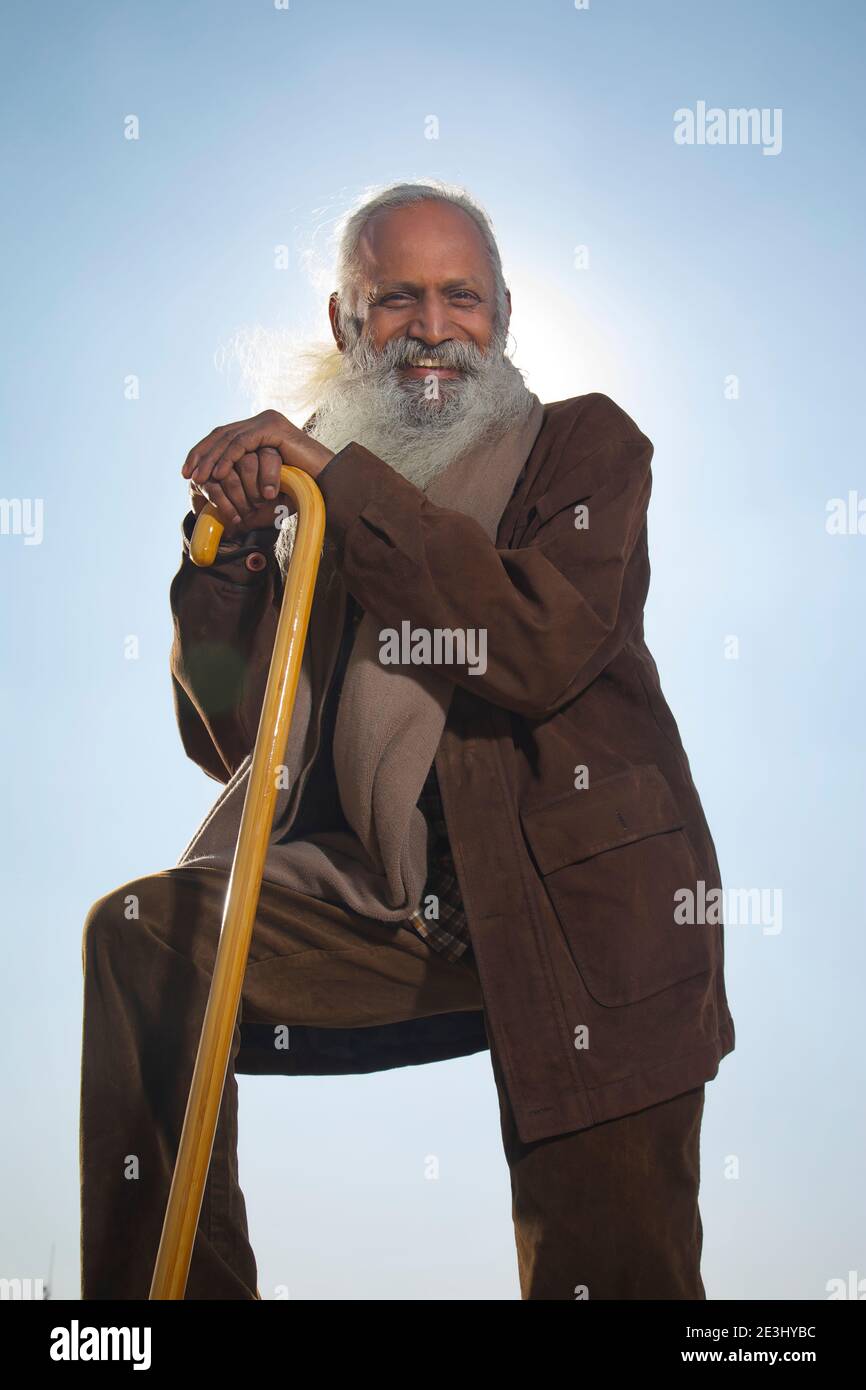 A BEARDED OLD MAN LOOKING AT CAMERA AND HAPPILY POSING WITH WALKING STICK Stock Photo