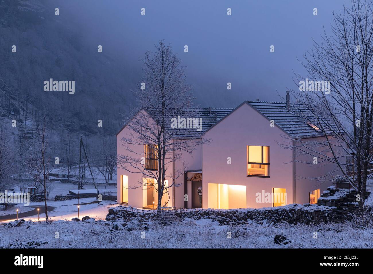 Modern white house with light windows surronded by mountain, snow and trees. Chirstmas mood. Nobody inside. Stock Photo
