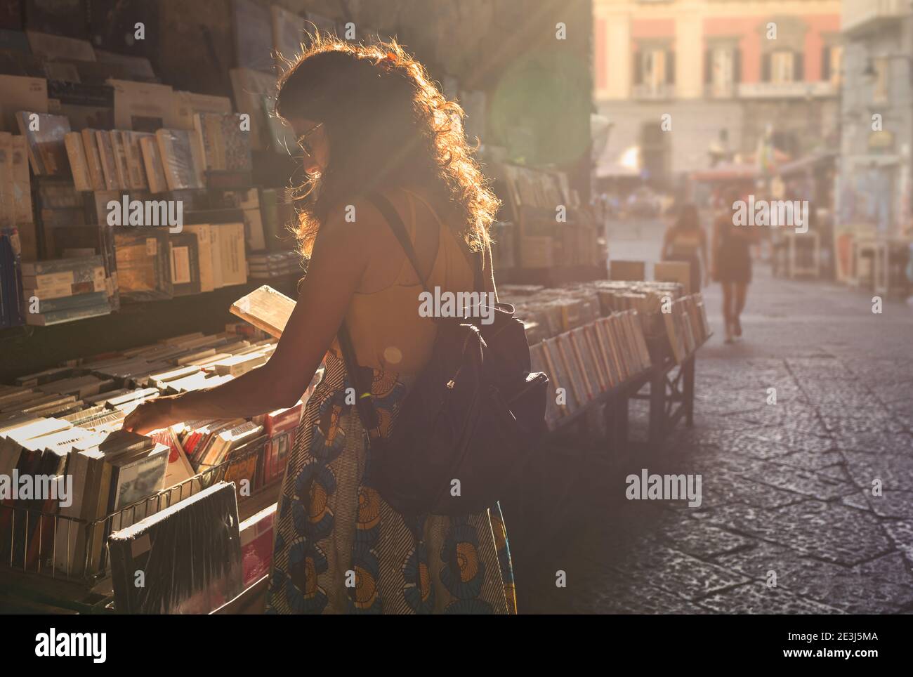 A young woman dressed in summer clothes chooses a book while shopping in an open air market in Europe. Stock Photo