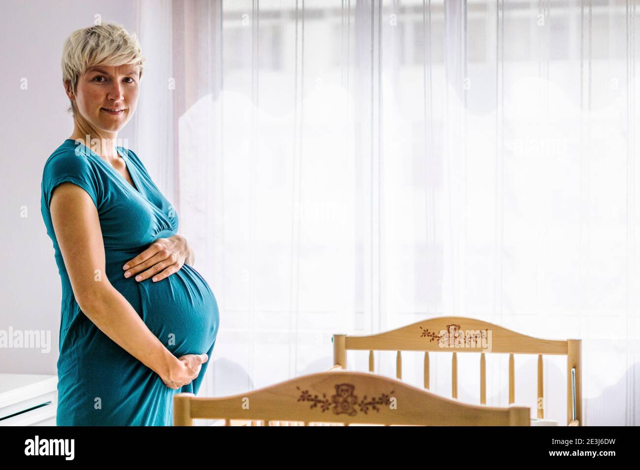 Pregnant woman holding her belly by baby cot next to the window Stock Photo