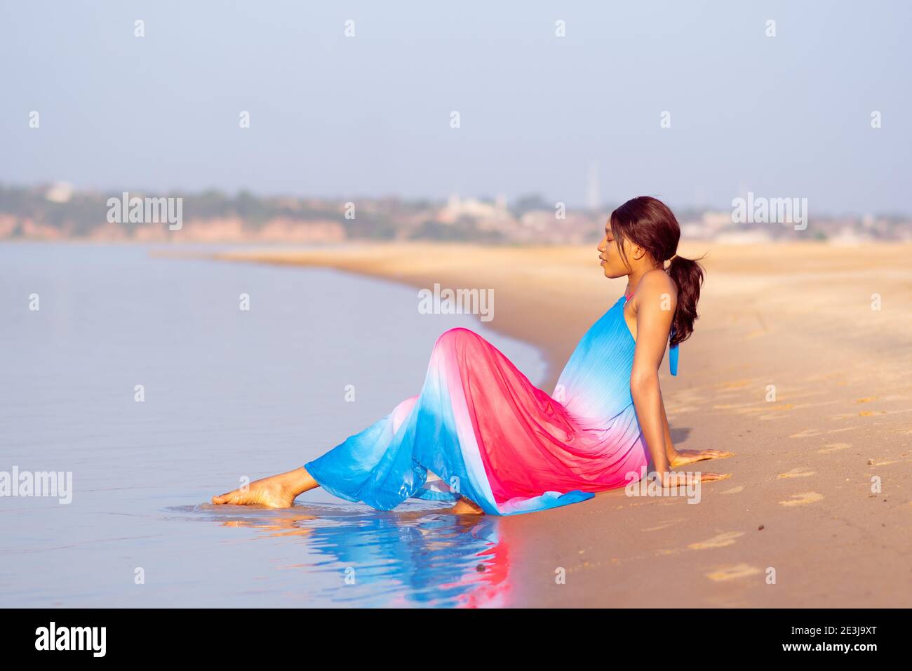 beautiful young african woman wearing a very colorful dress sitting on a beach alone and playing with the water with her feet Stock Photo