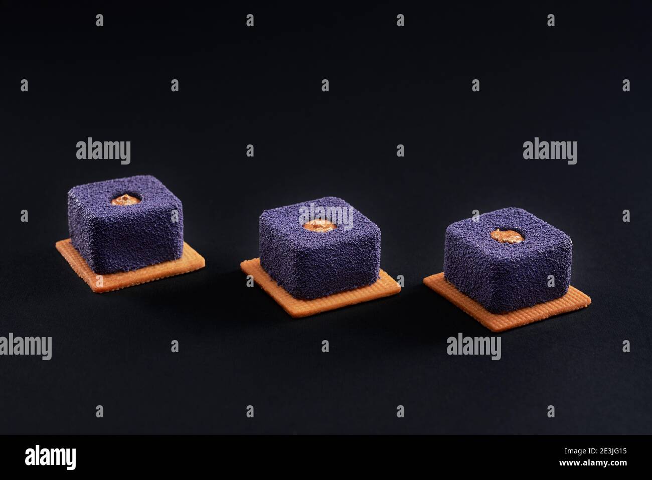 Closeup view of matte purple dessert filled with brown creme isolated on black studio background. Three small square matte cakes in row on cookies in Stock Photo