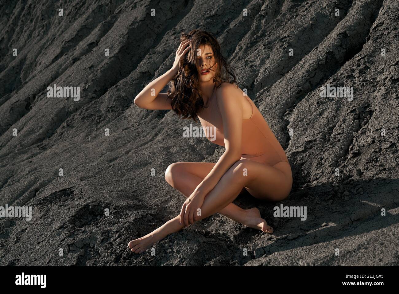 Side view of astonishing female model wearing beige body posing in dry empty quarry. Young barefoot stylish woman sitting on black sand outdoors in ho Stock Photo