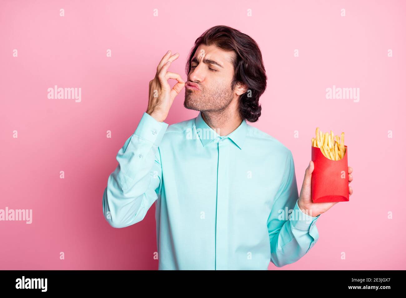 Photo portrait of man holding french fries in hand giving chef's kiss isolated on pastel pink colored background Stock Photo