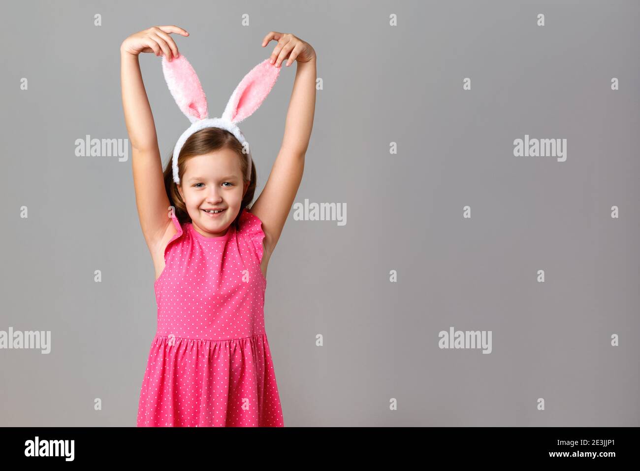 Happy easter. Cheerful little girl in a pink dress with polka dots on a gray background. The child holds his hands behind the rabbit ears. Stock Photo