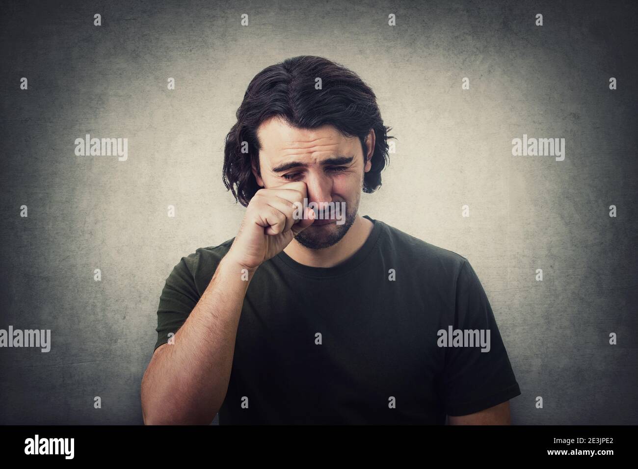 Portrait of desperate young man crying and wiping the tears with his hand isolated on grey wall background. Dissatisfied and disappointed guy sobbing Stock Photo