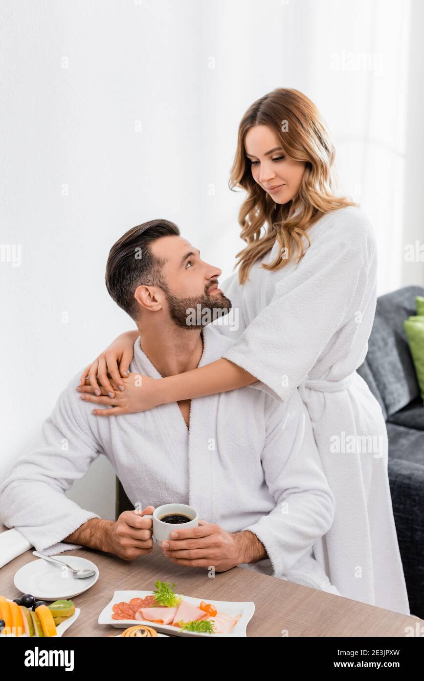 Woman in bathrobe hugging boyfriend with cup of coffee near breakfast on blurred foreground in hotel Stock Photo